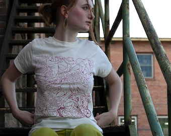 May include: A woman wearing a white t-shirt with a pink graphic print of a group of people. She is sitting on a set of stairs with her legs crossed. The t-shirt has the text "Tous Liberate" on it.