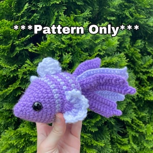 Op de afbeelding: Een gehaakte paarse en witte vis met een zwart oog. De vis wordt in een hand gehouden tegen een groene achtergrond.