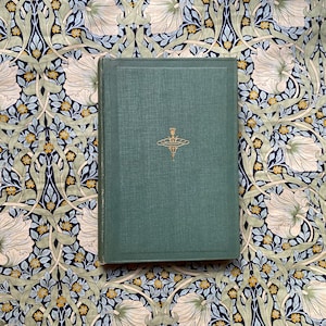 Puede incluir: Un libro de tapa dura verde con un diseño dorado en la portada. El libro está sobre una tela estampada con flores blancas y hojas verdes.