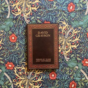 May include: A hardcover book titled "David Grayson: Essays of To-day and Yesterday" with a brown cover and gold lettering. The book is set against a backdrop of a floral patterned fabric in shades of blue, green, yellow, and red.