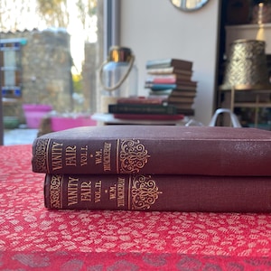 Op de afbeelding: Twee bordeauxrode antieke boeken met de titel "Vanity Fair" van W.M. Thackeray. De boeken liggen op een rood oppervlak met patronen. Gouden letters en decoratieve elementen sieren de ruggen.