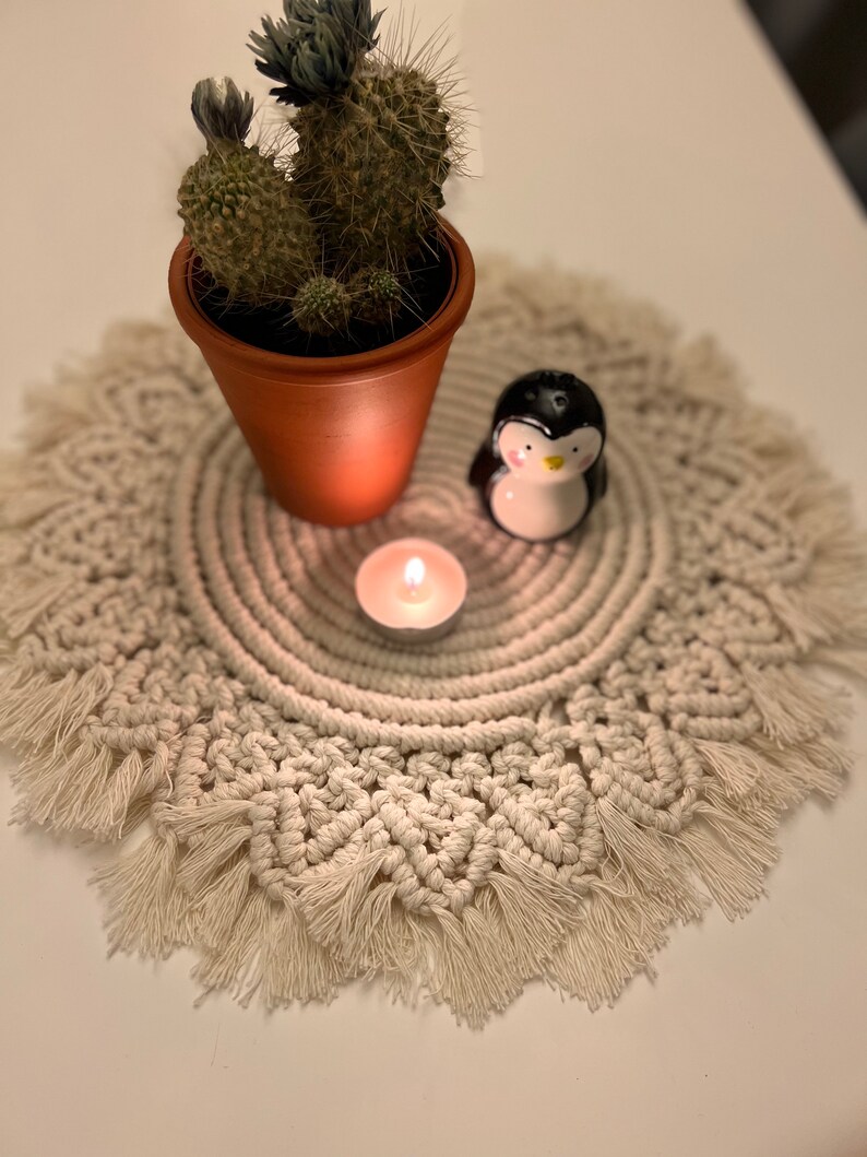 K&ouml;nnte beinhalten: Eine wei&szlig;e geh&auml;kelte Tischunterlage mit Fransenrand, ein kleiner Topf mit Kaktus, eine Keramik-Pinguinfigur und ein brennendes Teelicht.