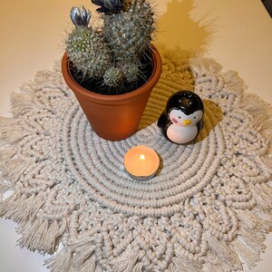 K&ouml;nnte beinhalten: Ein wei&szlig;er Makramee-Tischl&auml;ufer mit Fransen, ein kleiner Topf mit einem bl&uuml;henden Kaktus, eine kleine wei&szlig;e Kerze und eine schwarze Pinguinfigur.