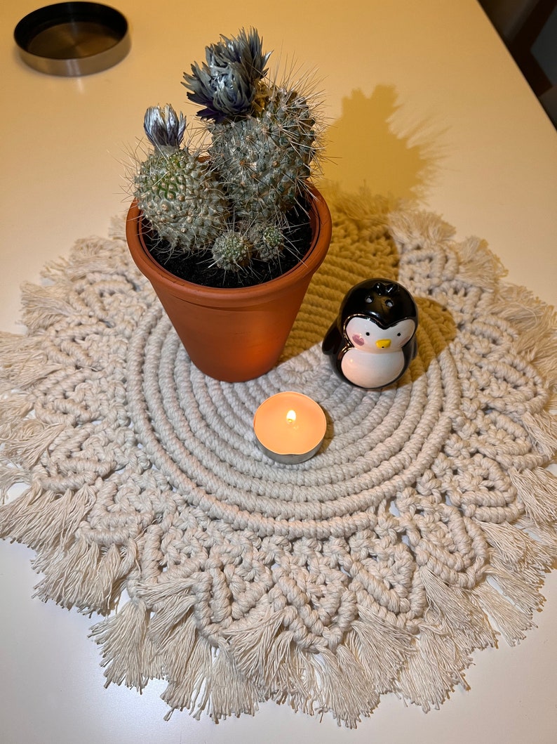 K&ouml;nnte beinhalten: Ein wei&szlig;er Makramee-Tischl&auml;ufer mit Fransen, ein kleiner Topf mit einem bl&uuml;henden Kaktus, eine kleine wei&szlig;e Kerze und eine schwarze Pinguinfigur.