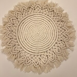K&ouml;nnte beinhalten: Ein runder, cremefarbener Makramee-Wandbehang mit Fransenrand.