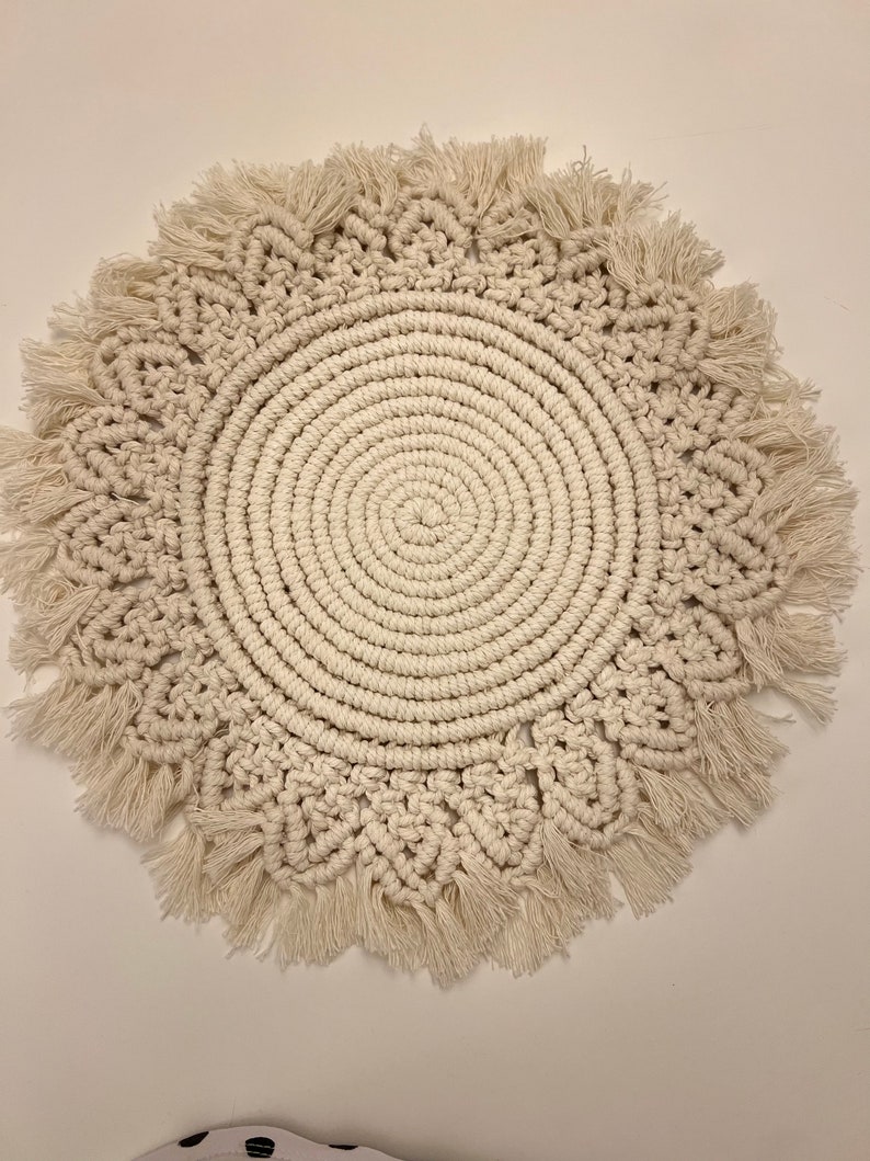 K&ouml;nnte beinhalten: Ein runder, cremefarbener Makramee-Wandbehang mit Fransenrand.