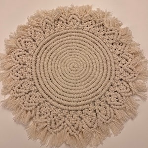 K&ouml;nnte beinhalten: Ein runder, cremefarbener Makramee-Untersetzer mit Fransenrand. Der Untersetzer ist aus Naturfasern gefertigt und hat ein kreisf&ouml;rmiges Design.