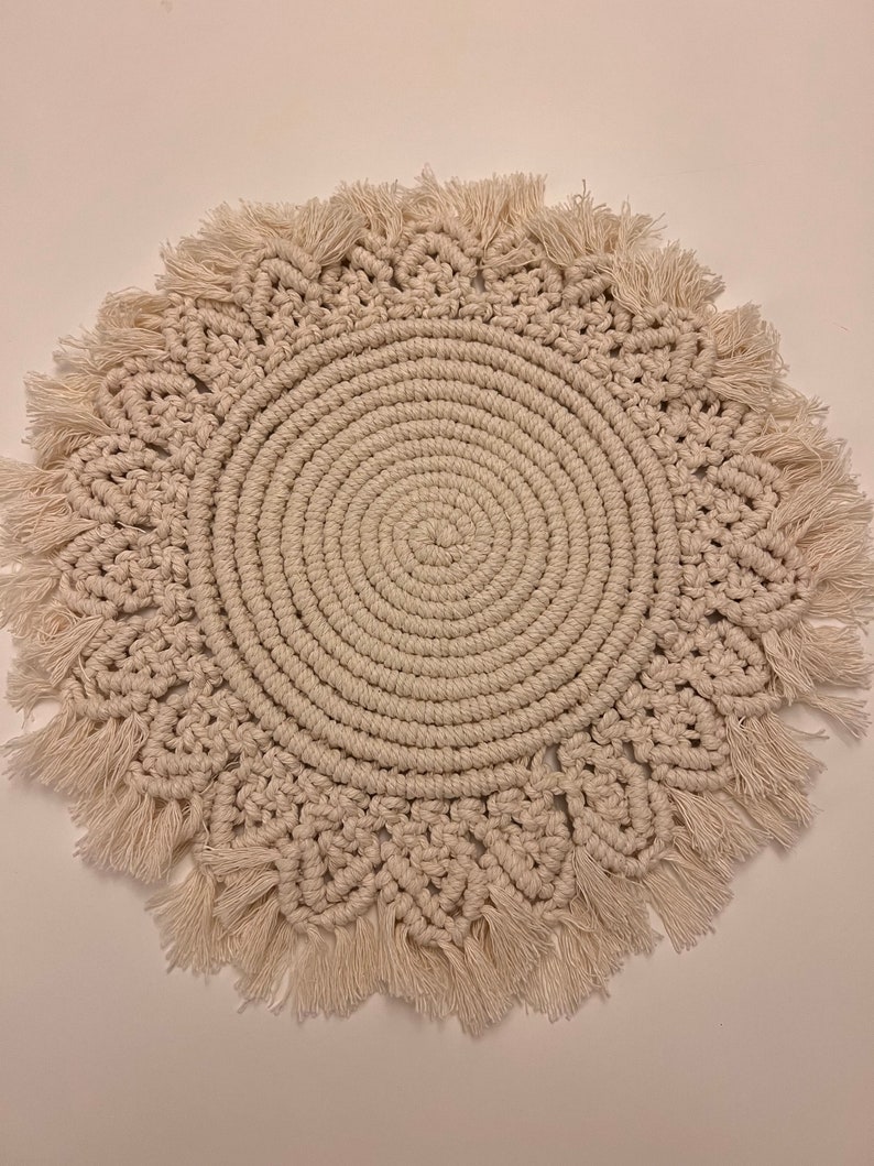 K&ouml;nnte beinhalten: Ein runder, cremefarbener Makramee-Untersetzer mit Fransenrand. Der Untersetzer ist aus Naturfasern gefertigt und hat ein kreisf&ouml;rmiges Design.