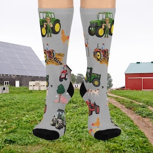 May include: A pair of gray socks with a pattern of tractors, farmers, chickens, and other farm-related imagery. The socks are shown on a model's feet against a backdrop of a farm scene.
