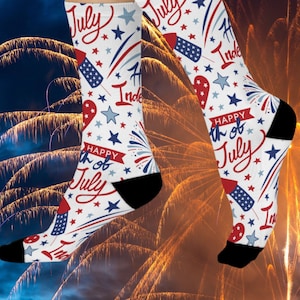 Puede incluir: Calcetines blancos con un diseño festivo del 4 de julio. Los calcetines presentan fuegos artificiales rojos, blancos y azules, estrellas y las palabras "Happy 4th of July" y "July". Los talones y las puntas son negros.