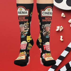 May include: Black socks with a cinema theme. The socks feature popcorn, film reels, movie tickets, and the words "Cinema" and "Movie Time." The socks have a red, white, and yellow colour scheme.