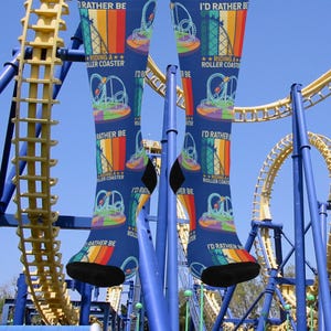May include: Blue knee-high socks with a roller coaster design. The socks feature a colorful rainbow design with the text "I'd Rather Be Riding a Roller Coaster". The socks are worn in front of a roller coaster.