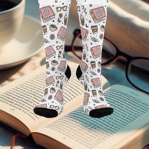 May include: White knee-high socks with a repeating pattern of pink books, coffee cups, eyeglasses, and flowers. The socks have black heels and toes. The socks are on a person's legs, with an open book in the foreground.