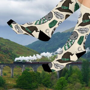 May include: Cream-colored socks with a repeating pattern of a wizard hat, feathers, an alien with a striped scarf, and other magical elements. The socks have black heels and toes. The background shows a train on a bridge in a green landscape.