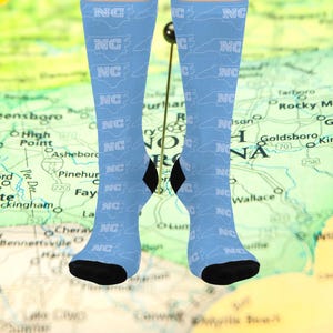 May include: Light blue socks with a repeating pattern of the letters "NC" and the outline of the state of North Carolina. The socks have black heels and toes. The background is a map of North Carolina.