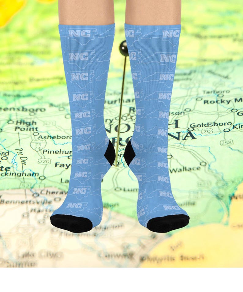 May include: Light blue socks with a repeating pattern of the letters "NC" and the outline of the state of North Carolina. The socks have black heels and toes. The background is a map of North Carolina.