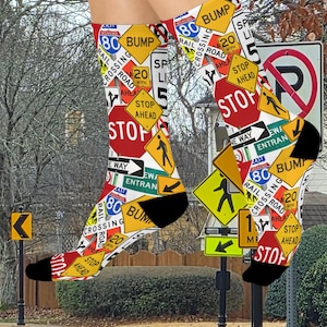 May include: A pair of novelty socks featuring a collage of road signs, including "STOP," "BUMP," and speed limit signs. The socks have black heels and toes, with a white background. The design includes signs with the text "RAILROAD CROSSING" and "ENTRAN".