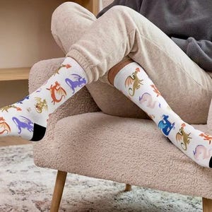 May include: A pair of white crew socks featuring a repeating pattern of small, colorful dragons in shades of orange, purple, blue, and green. The socks have solid black toe and heel sections, providing a distinct contrast to the white fabric.