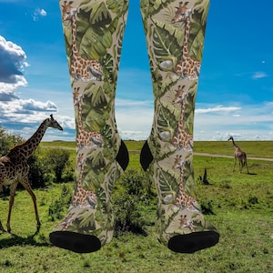 May include: A pair of knee-high socks featuring a repeating pattern of giraffes and tropical leaves in shades of green, tan, and brown. The socks have black heels and toes. The background shows a savanna landscape with giraffes and a blue sky.