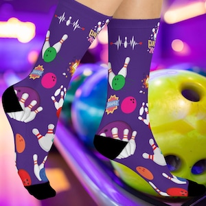 May include: Purple socks with a bowling theme. The socks feature bowling pins, bowling balls, and a heartbeat graphic. The socks have black heels and toes. The socks are on a person's legs.