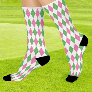 May include: A pair of socks with a white background and a repeating pattern of green and pink diamonds. The heel and toe of each sock are black. The socks are worn on a person's legs, with a green background.