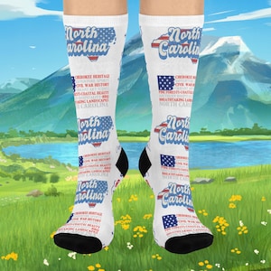 May include: White socks with black heels and toes, featuring a North Carolina design. The design includes the state outline, the words "North Carolina," and text about the state's heritage, sports, and landscapes. The socks are displayed against a scenic background.