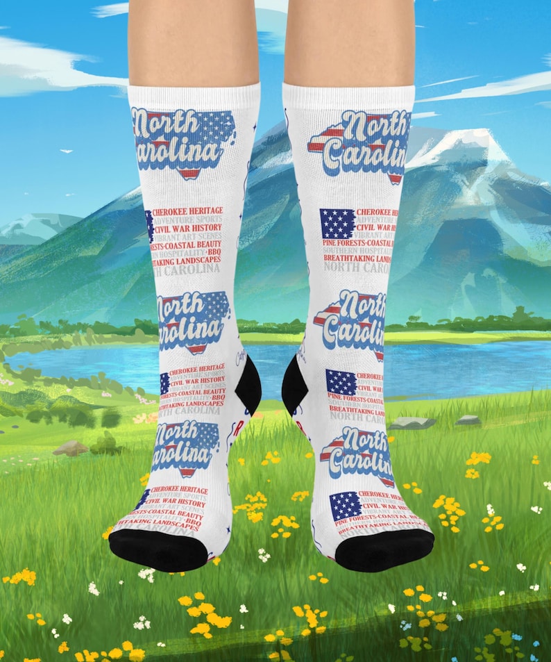 May include: White socks with black heels and toes, featuring a North Carolina design. The design includes the state outline, the words "North Carolina," and text about the state's heritage, sports, and landscapes. The socks are displayed against a scenic background.