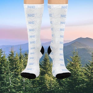 K&ouml;nnte beinhalten: Wei&szlig;e Socken mit einem sich wiederholenden Muster aus "NC" und der Umrisslinie von North Carolina in Hellblau. Die Fersen und Zehen sind schwarz. Der Hintergrund zeigt eine Bergkette und immergr&uuml;ne B&auml;ume.