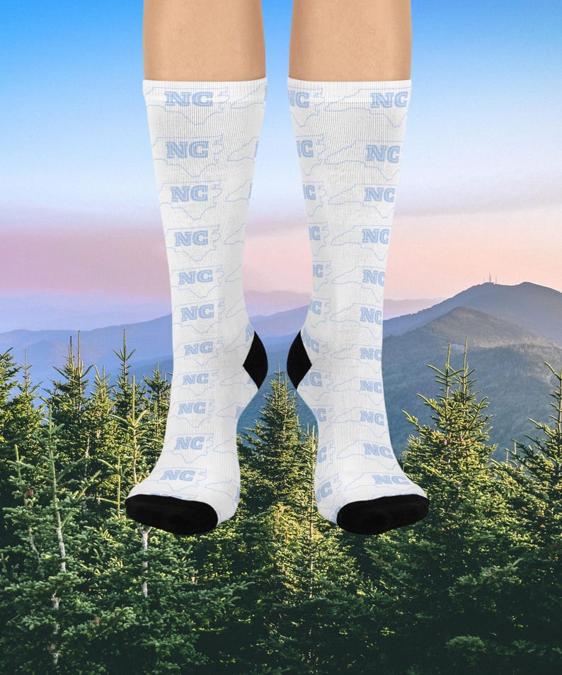 K&ouml;nnte beinhalten: Wei&szlig;e Socken mit einem sich wiederholenden Muster aus "NC" und der Umrisslinie von North Carolina in Hellblau. Die Fersen und Zehen sind schwarz. Der Hintergrund zeigt eine Bergkette und immergr&uuml;ne B&auml;ume.