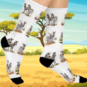 May include: White socks with a black heel and toe feature a repeating pattern of zebras in a savanna setting. The socks have a colorful illustration of zebras in a natural habitat, with a background of trees and grass.