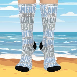 May include: A pair of knee-high socks with a light blue and white design. The socks feature text including "America", "Raleigh", "Carolina", and "University". The toes and heels are black. The background shows a beach scene.