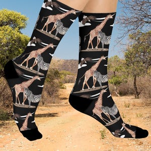May include: Black socks with a repeating pattern of giraffes, zebras, and eagles. The giraffes and zebras are on surfboards. The socks have black heels and toes. The background shows a dirt path and trees.
