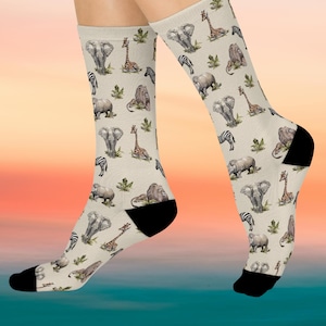 May include: A pair of beige socks featuring a repeating pattern of watercolor-style African animals, including elephants, giraffes, zebras, lions, and rhinos. The socks have black heels and toes, and the animals are set against a backdrop of green foliage.