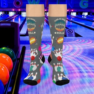 May include: Gray knee-high socks with a bowling theme. The socks feature bowling pins, bowling balls, and the phrases "This is how I roll" and "Strike!" in a colorful design. The background shows a bowling alley.