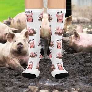 May include: White socks with a repeating pattern of cartoon pigs, mud splashes, and the word "oink!" in black. The socks have black heels and toes. The background shows pigs in a muddy pen.