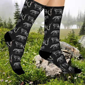 May include: Black socks featuring a repeating pattern of white wolves and curved tusks. The socks have black heels and toes. The socks are knee-high and are worn outdoors in a grassy area.