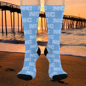 May include: Light blue knee-high socks with the white letters "NC" repeated in a pattern. The socks have black heels and toes. The background shows a beach scene with a pier and a sunset.