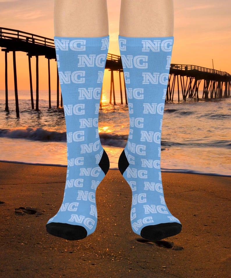 May include: Light blue knee-high socks with the white letters "NC" repeated in a pattern. The socks have black heels and toes. The background shows a beach scene with a pier and a sunset.