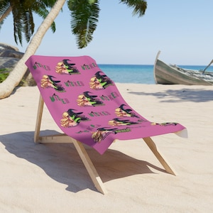 Könnte beinhalten: Ein pinkfarbener Strandstuhl mit einem Handtuch mit einem Cartoon-Hexen-Design. Die Hexe hat blondes Haar, ein grünes Gesicht und einen schwarzen Hut. Das Wort "WICKED" ist auf das Handtuch gedruckt. Der Stuhl ist aus Holz.