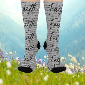 May include: A pair of gray socks with the word "Faith" in a cursive font. The socks have a black heel and toe. The background is a light blue sky with a field of flowers.