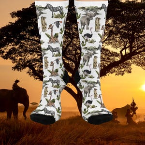 May include: White socks with a repeating pattern of African animals including zebras, giraffes, elephants, and ostriches. The socks have black heels and toes. The background shows a sunset over a savanna.
