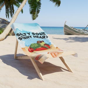 Op de afbeelding: Een strandstoel met een "Salty Soul Sunny Heart" ontwerp. De stoel heeft een cartoon schildpad met een zonnebril die op een rode band drijft. De achtergrond toont een strand, oceaan en een boot.