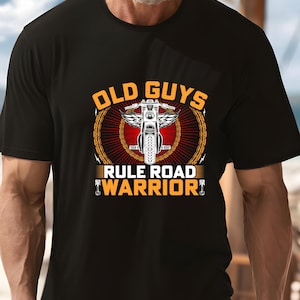 Puede incluir: Camiseta negra con un gráfico de motocicleta y el texto "OLD GUYS RULE ROAD WARRIOR". El diseño presenta una motocicleta con alas, un borde de cadena y una combinación de colores rojo y dorado.