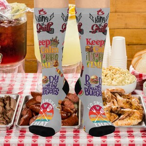 May include: Gray knee-high socks with barbecue-themed graphics. The socks feature text like "King of Grill", "Keep Calm It's BBQ Time", and "I'd Smoke That". The design includes images of burgers, crowns, and grilling tools.