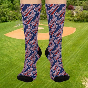 Könnte beinhalten: Marineblaue Baseballsocken mit einem sich wiederholenden Muster des Wortes "Baseball" in Weiß und Rot. Die Socken haben schwarze Fersen und Zehen. Der Hintergrund zeigt ein Baseballfeld. Sportliches Accessoire.