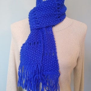 May include: A blue knitted scarf with fringe on the ends. The scarf is draped over a beige sweater.