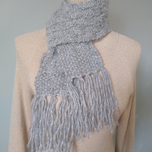May include: A silver grey knitted scarf with fringe on a beige sweater.