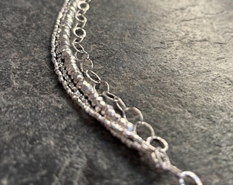 May include: A multi-strand silver chain necklace featuring a combination of delicate textured bead links and smooth oval links. The metallic silver finish reflects light across the various chain styles, creating a layered appearance with distinct link patterns.