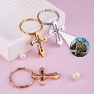 May include: Three silver and gold cross keychains with a small clear stone in the center of each cross. The keychains are on a pink surface with white pearls.