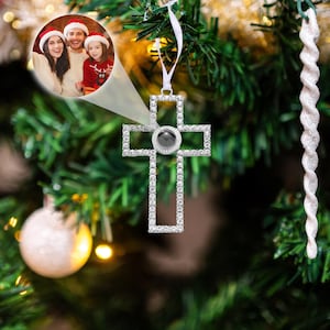 May include: A silver cross-shaped Christmas ornament with clear crystals and a dark gray center stone. The ornament hangs from a white ribbon on a green Christmas tree. A family photo is in the background.